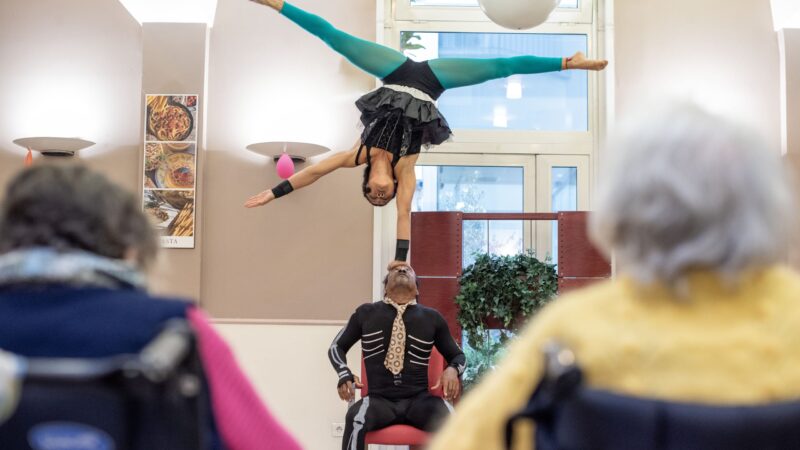 Un duo d’acrobates en équilibre sur scène, devant des résidents assis qui applaudissent.