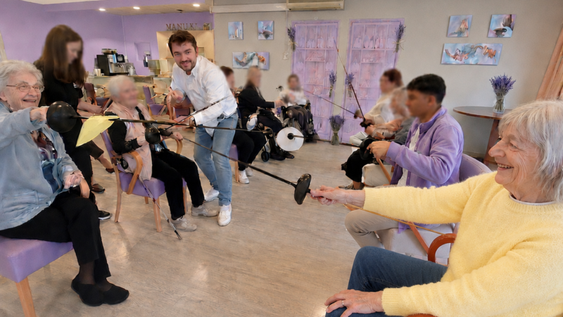 Des résidents en fauteuil pratiquent l’escrime souple avec des bâtons en mousse, encadrés par un animateur.
